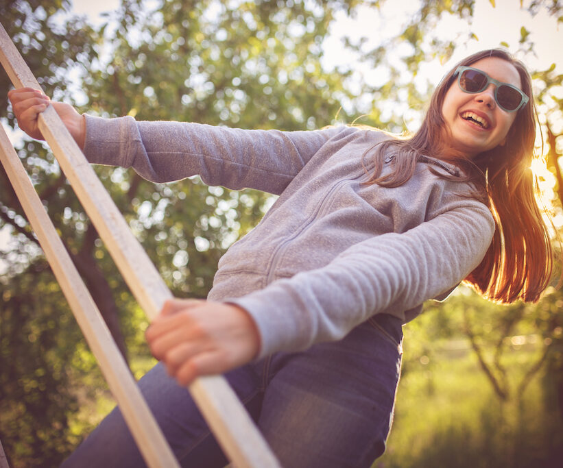 girl happy smiling outdoors, playing on playground with sunglasses on and woods behind
