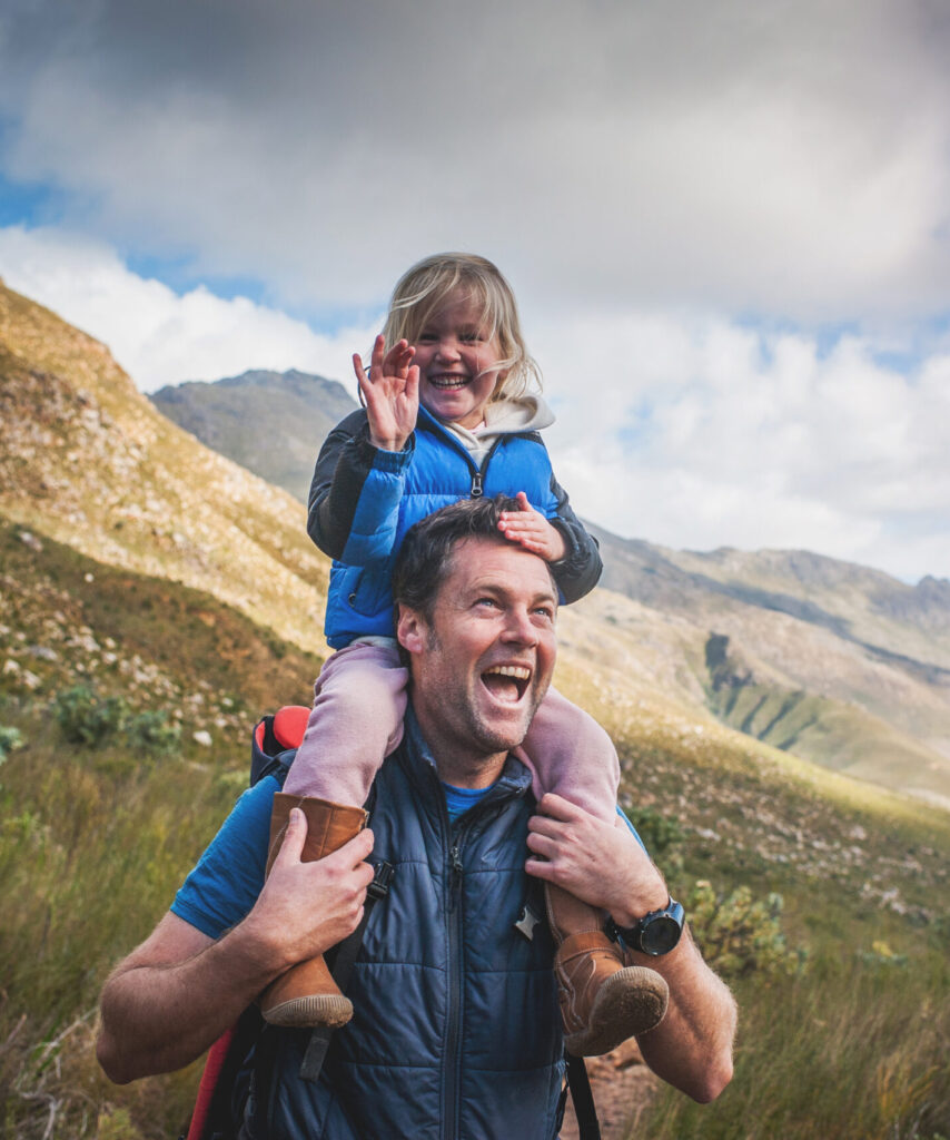 Father and daughter smiling and laughing on trail with daughter waving at the camera and mountain hills behind
