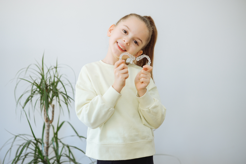 Young brunette child in front of blank wall holding Invisalign clear aligners, Invisalign First at GNW Orthodontics